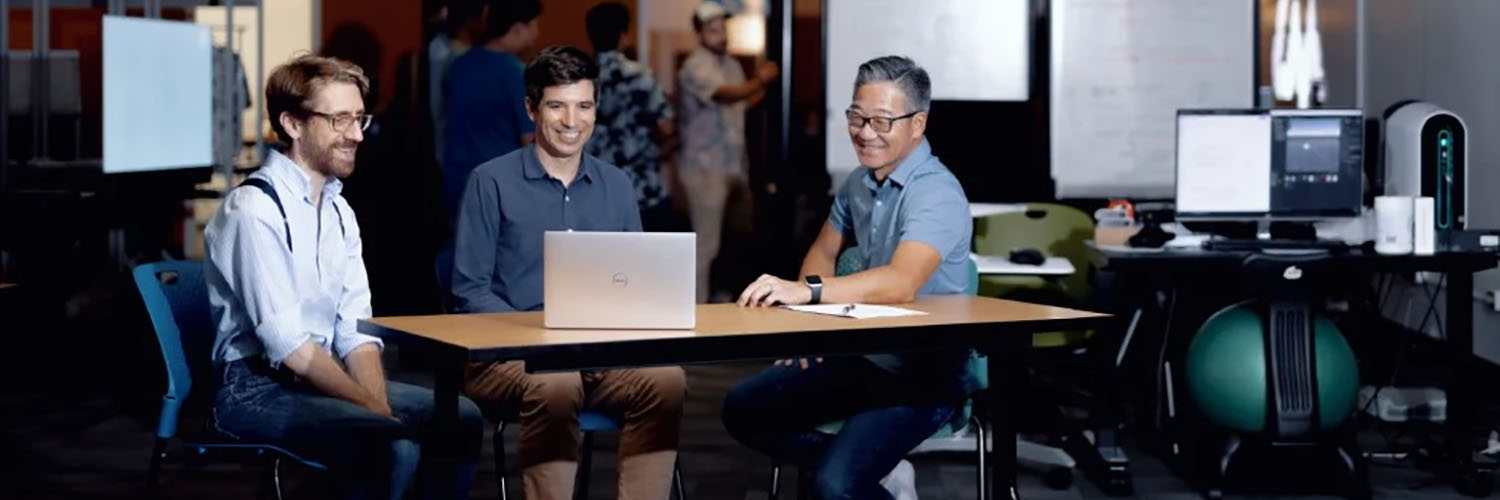 Three Guys Sitting At A Table Around A Laptop